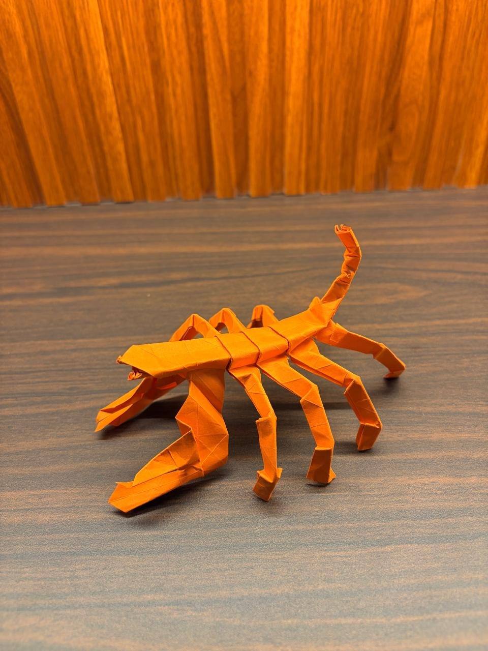 An orange paper scorpion on a dark desk, pincers forward and its tail curling up behind it.