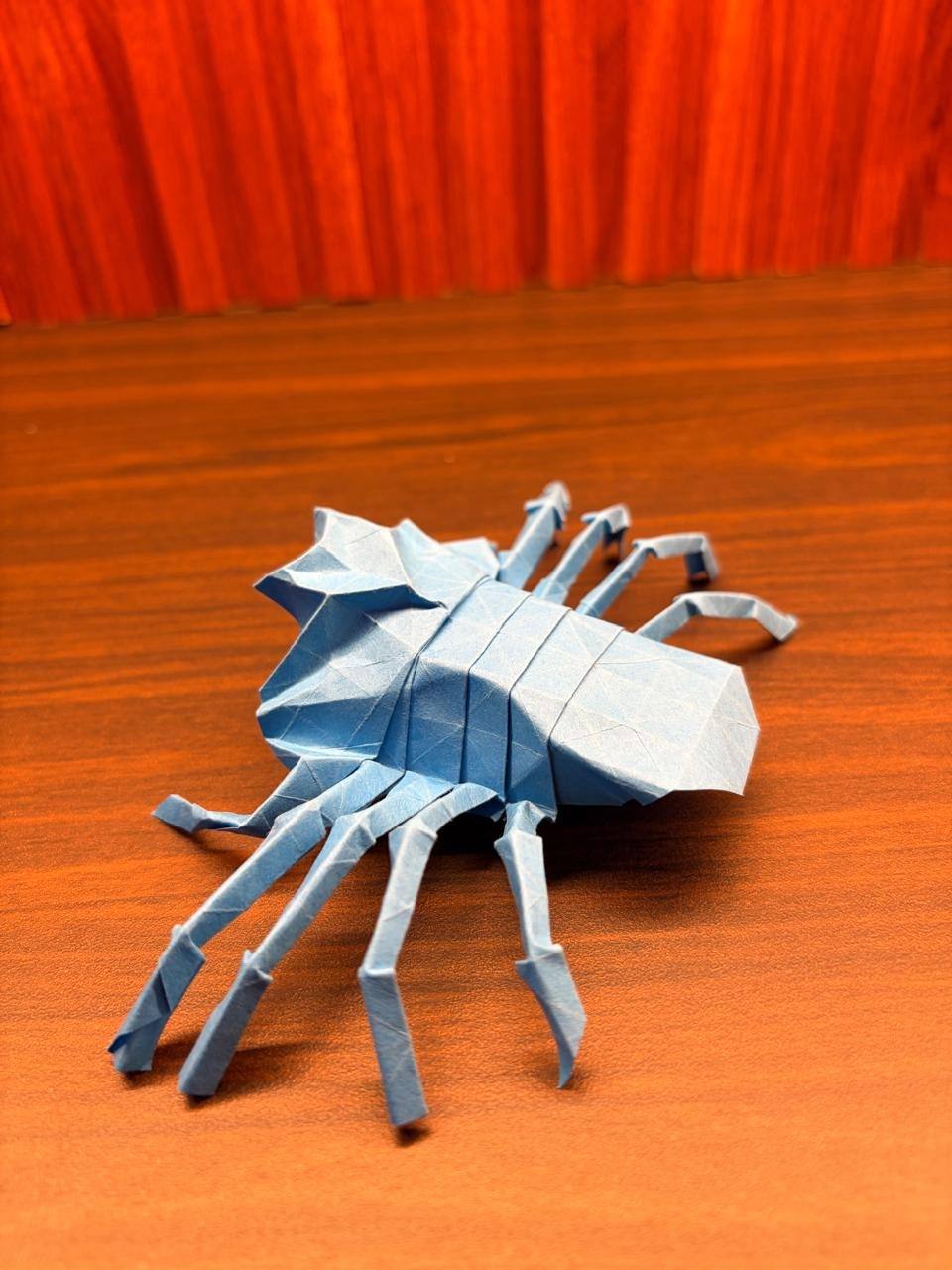 A light blue paper spider photographed from above, its eight folded legs spread across an orange desk.
