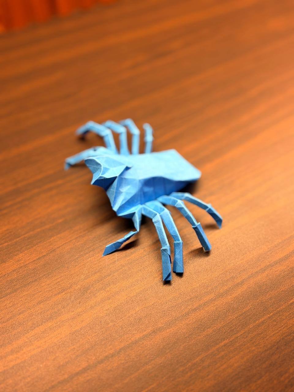 The blue paper spider low in the frame, its front legs sharp against a blurred wooden desk.