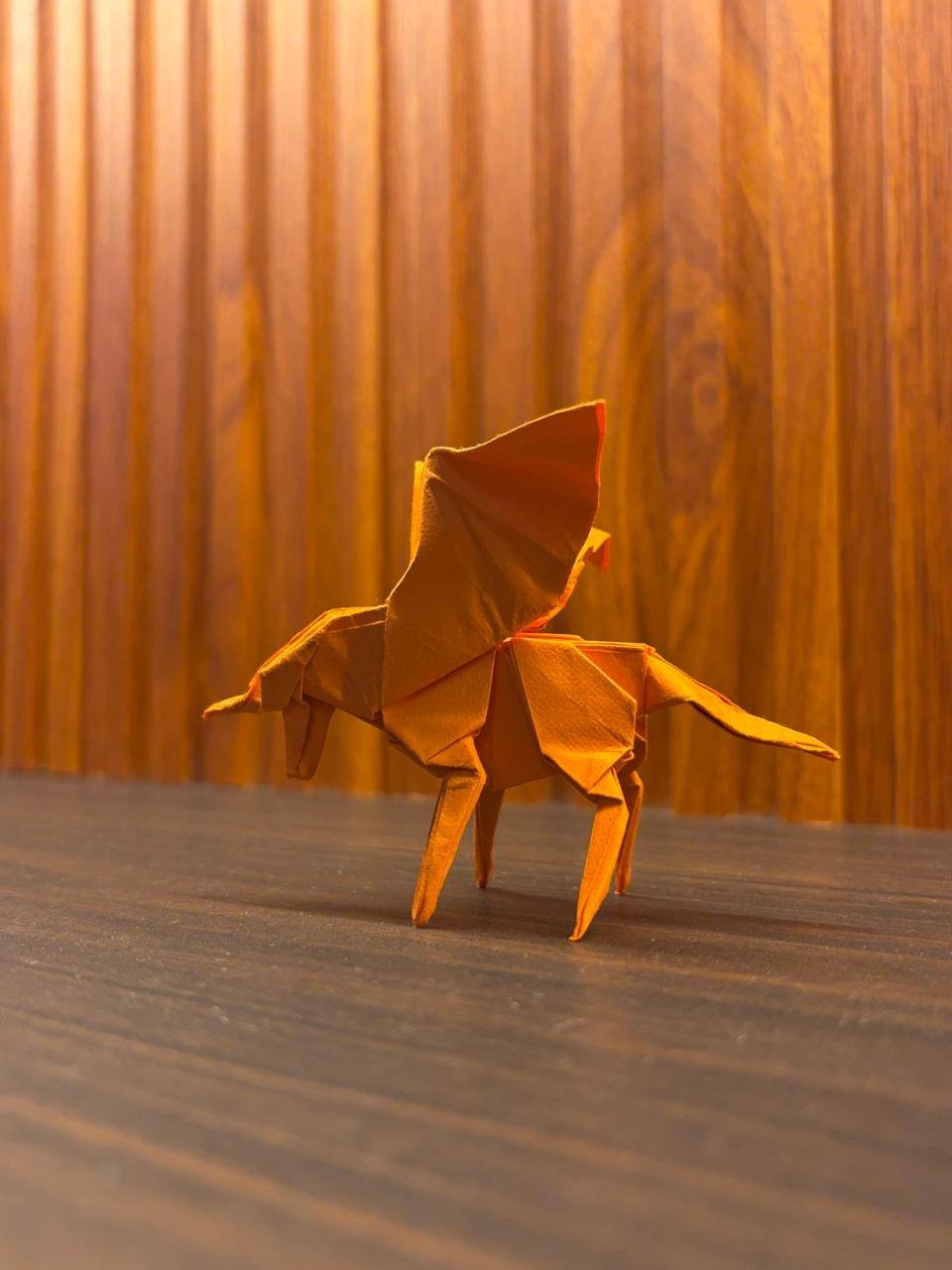 An orange paper Pegasus standing on four legs with its wings raised, in front of warm wooden panels.